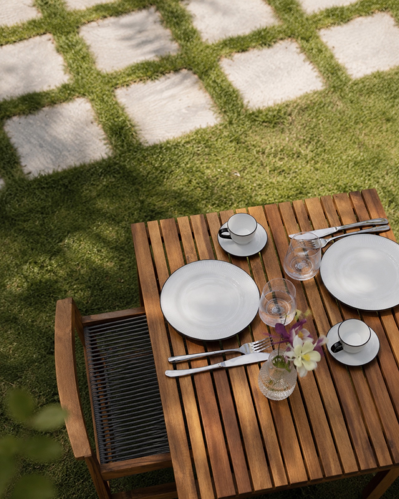 A wooden garden table set with white plates, wine glasses and a small vase of garden flowers, on grass and stepping stones