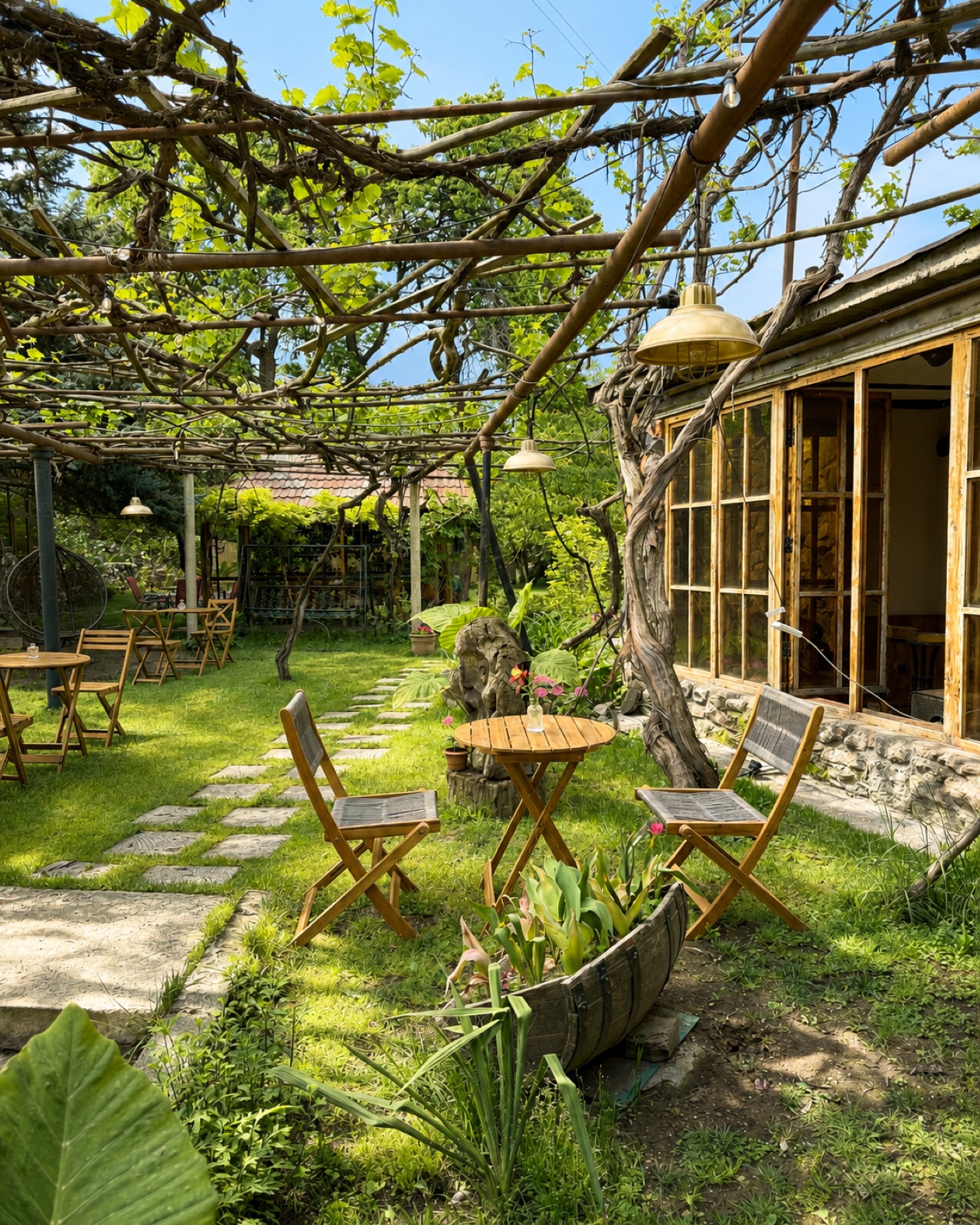 The walled garden in daylight, wooden cafe tables and chairs under a vine pergola, stepping stones across the grass, the house on the right
